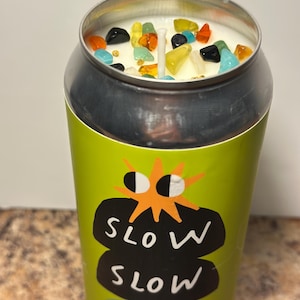 May include: A candle in a repurposed aluminum can. The can is green with the words "SLOW SLOW" in a black bubble design. The candle wax is white, topped with colorful decorative stones.