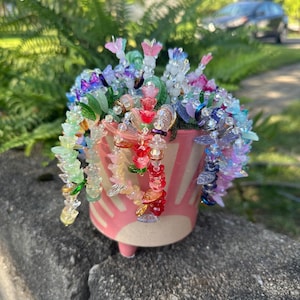 May include: A pink ceramic pot with a white stripe design, filled with colorful beaded flowers. The flowers are in shades of pink, red, blue, green, and yellow. The pot is sitting on a gray stone surface.