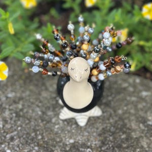 May include: A ceramic penguin-shaped vase, black and white, with a cream-coloured face and closed eyes. The vase holds beaded stems in various colours, including brown, grey, and cream. The vase is set on a textured grey surface, with blurred greenery in the background.