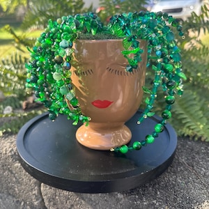 May include: A brown ceramic face planter with closed eyes and red lips, adorned with cascading green beaded strands and various green beads. The planter sits on a black round tray, creating a unique decorative piece.
