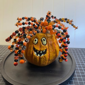 May include: A textured orange pumpkin figurine with a carved smiling face featuring white eyes and jagged teeth. The top of the pumpkin is adorned with numerous wire strands decorated with orange, black, and clear beads in various shapes.