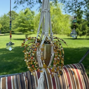 May include: A handmade macrame plant hanger with a glass container filled with plants and adorned with amber and yellow beads. The plant hanger is suspended outdoors, with a beaded chain and a crystal prism hanging nearby.