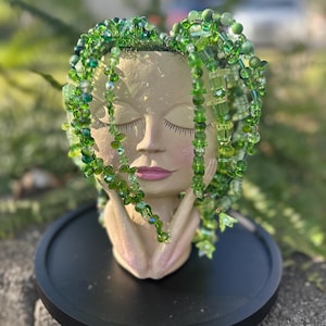 Puede incluir: Una maceta decorativa con forma de rostro sereno con los ojos cerrados, adornada con cuentas y cristales verdes en cascada, que se asemejan a cabello fluido. La maceta descansa sobre una base circular negra, creando una exhibición caprichosa y artística.