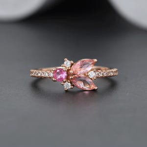 May include: A rose gold ring featuring a cluster of pink gemstones and small white stones. The ring's band is encrusted with small, clear stones. The central design includes a round pink stone and two marquise-shaped pink stones.