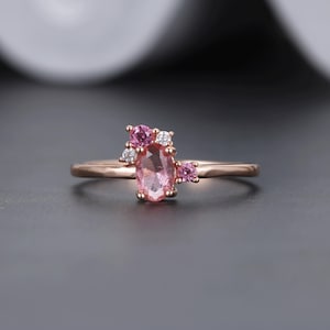 May include: Rose gold ring featuring a large oval pink gemstone, surrounded by smaller pink and white gemstones. The ring has a delicate band and a cluster design, perfect for a special occasion.