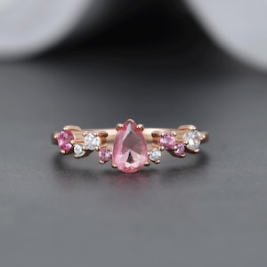 May include: A rose gold ring with a central pear-cut pink gemstone, flanked by smaller pink and white stones. The ring's design is delicate and floral, suitable for a special event. The ring's design is delicate and floral.