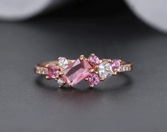 May include: Rose gold ring featuring a large rectangular pink gemstone surrounded by smaller pink and white stones. The band is adorned with tiny clear stones, creating a cluster design. The ring is set against a dark gray background.