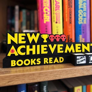Op de afbeelding: Een zwart bord met gele tekst NEW ACHIEVEMENT! BOOKS READ, voorzien van een geel trofee-icoon, drie rode schedels en een rood oog. Daarnaast staat een zwarte teller met drie blokjes die GLURP, GLURP!, een boekicoon en het cijfer 1 weergeeft.
