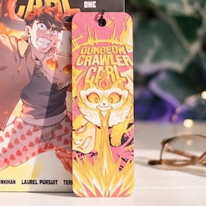 May include: A graphic novel titled Dungeon Crawler Carl Volume One featuring a character in heart-patterned boxers, accompanied by a matching rectangular bookmark with a cat illustration, both displaying the Dungeon Crawler Carl logo text.