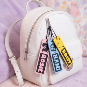 Op de afbeelding: Een witte leren rugzak met drie rechthoekige tashangers aan de ritssluiting. De hangers zijn roze, blauw en geel en tonen elk een stripfiguur en de namen ROSIE, NAMI en DAISY in een vetgedrukte zwarte lettertype op de voorzijde.