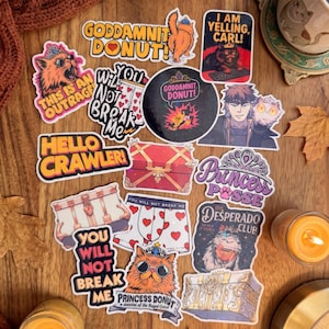 May include: A collection of assorted stickers featuring various illustrations including cats, treasure chests, and boxers with heart patterns. Text includes phrases like GODDAMNIT DONUT, I AM YELLING CARL, HELLO CRAWLER, YOU WILL NOT BREAK ME, PRINCESS POSSE, and THE DESPERADO CLUB.