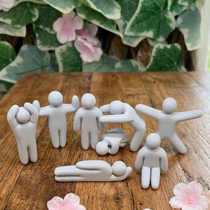 Op de afbeelding: Een verzameling van zeven kleine, mat lichtgrijze beeldjes in de vorm van gestileerde menselijke figuren in verschillende houdingen, waaronder staand, rekkend en liggend, gerangschikt op een houten oppervlak met verspreide lichtroze bloemblaadjes.