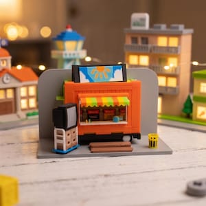 Op de afbeelding: Een miniatuur oranje foodtruck bouwset met een geel en groen gestreepte luifel, een bord met een zongrafiek, een kleine zwarte televisie op een wit karretje en een gele prullenbak, alles gemonteerd op een grijze displaybasis.