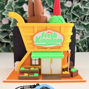 Può includere: Un negozio di alimentari in miniatura stampato in 3D a forma di carrello della spesa giallo con un'insegna con la scritta Fresh Kingdom. La struttura presenta una tenda verde, porte bianche e scaffali pieni di piccole cassette di prodotti. È incluso un portachiavi blu.