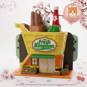 Op de afbeelding: Een gele en groene miniatuur supermarkt met de tekst "Fresh Kingdom". Het bevat miniatuur voedsel zoals brood, ketchup en sla. Kersenbloesemtakken omlijsten de scène. Het model is gedetailleerd en kleurrijk.