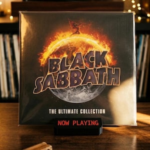 Op de afbeelding: Een Black Sabbath The Ultimate Collection vinyl platenhoes met een brandende planeetgrafiek en een kleine gevleugelde figuur. De plaat staat op een zwarte standaard met de rode tekst NOW PLAYING gedrukt op de voorzijde van de voet.