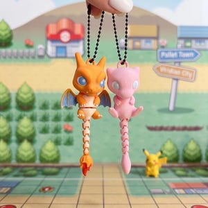 May include: Two articulated plastic keychains featuring orange and pink creature figures with long, segmented tails. The orange figure has blue wings and large eyes, while the pink figure has a smooth, rounded head. Both hang from black ball chains.