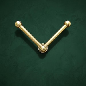 Puede incluir: Un gancho de pared en forma de V con dos brazos de latón pulido que se extienden desde una base circular central. Cada brazo es cilíndrico con un acabado liso y termina en un extremo esférico redondeado, creando un diseño simétrico.