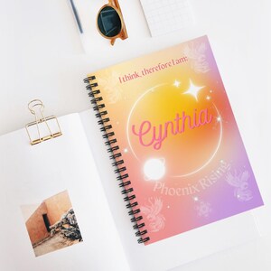 May include: Spiral-bound notebook with a colorful cover featuring the name "Cynthia" in pink script. The cover has a gradient background of orange, yellow, and purple with the text "I think, therefore I am: Phoenix Rising."