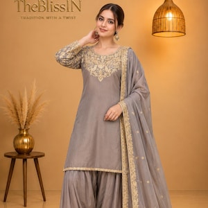 Puede incluir: Un conjunto de salwar kameez gris que incluye una túnica de manga larga con bordados dorados alrededor del cuello, pantalones plisados a juego con dobladillos bordados en oro y una dupatta gris transparente con borde dorado.