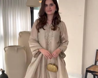May include: A beige silk dress with embroidered detailing and flared sleeves. The dress features a contrasting burgundy hem with white floral embroidery. A small gold clutch is held in front.