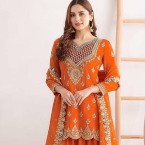 Könnte beinhalten: Ein orangefarbenes, besticktes Outfit mit passendem Schal. Das Ensemble weist aufwendige Gold- und Silberdetails am Ausschnitt, an den Ärmeln und am Saum auf. Das Outfit ist im traditionellen Stil gehalten, wahrscheinlich für besondere Anlässe.