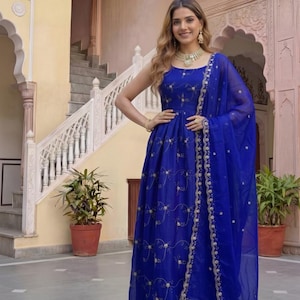 May include: A sleeveless, royal blue gown with gold embroidery and a matching sheer scarf. The dress features a fitted bodice and a flowing skirt. The model is wearing gold jewelry. The background includes a staircase and potted plants.
