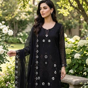 Könnte beinhalten: Schwarzes zweiteiliges Outfit mit Stickerei. Das langärmlige Oberteil und die passende Hose weisen aufwendige weiße Blumenstickereien auf. Ein durchsichtiger schwarzer Schal mit gewelltem Rand vervollständigt das Ensemble. Das Outfit wird im Freien in einem Garten getragen.