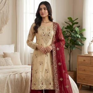Puede incluir: Un conjunto salwar kameez beige bordado con un dupatta granate. La t&uacute;nica de manga larga y los pantalones de pernera ancha est&aacute;n adornados con bordados dorados. El dupatta granate presenta un borde bordado a juego.