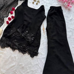 May include: A black two-piece outfit featuring a long-sleeved top with floral lace detailing and flared pants. The top has a square neckline. The outfit is displayed on a white, textured surface, with a black sheer scarf and decorative accessories.