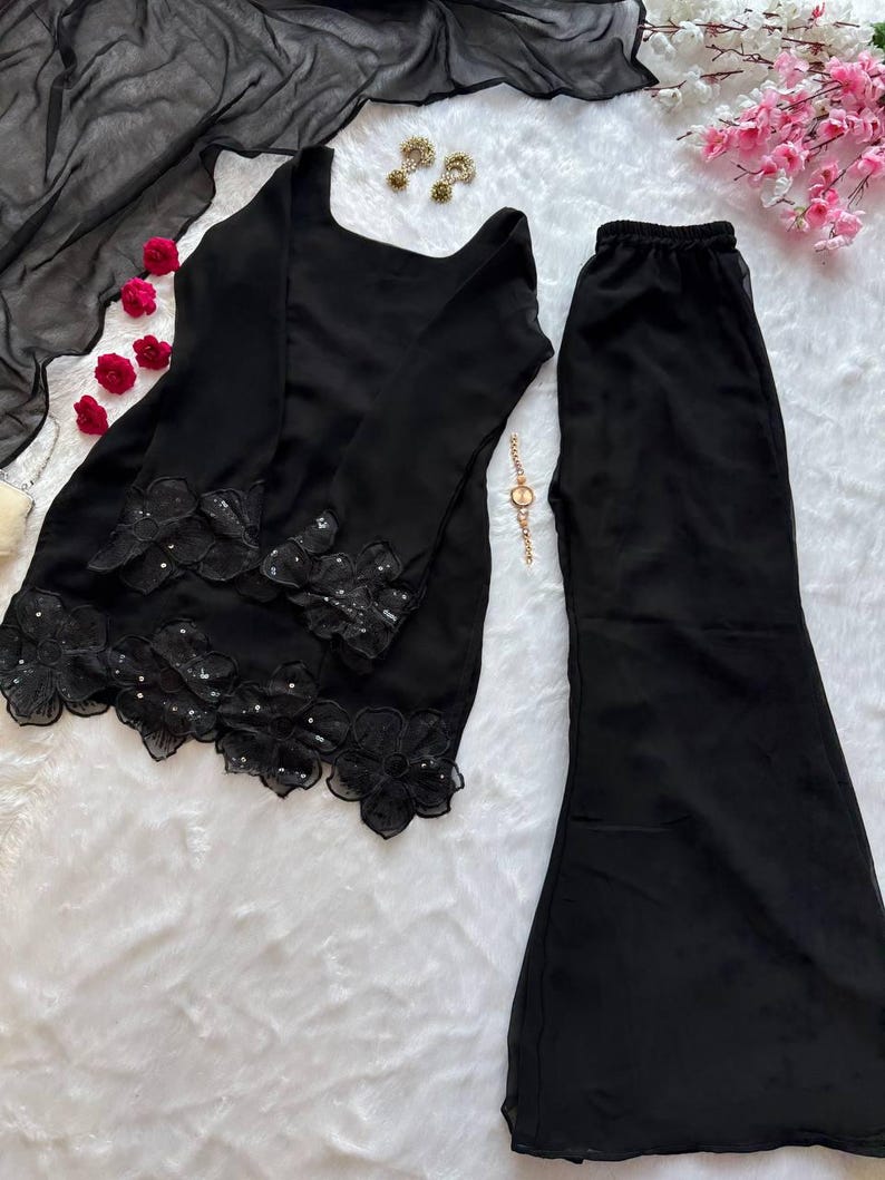 May include: A black two-piece outfit featuring a long-sleeved top with floral lace detailing and flared pants. The top has a square neckline. The outfit is displayed on a white, textured surface, with a black sheer scarf and decorative accessories.
