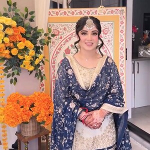 May include: A woman in a navy blue and gold embroidered outfit, including a headpiece, stands in front of a decorative backdrop. The outfit features floral patterns and intricate detailing. Floral arrangements in yellow and orange are visible.