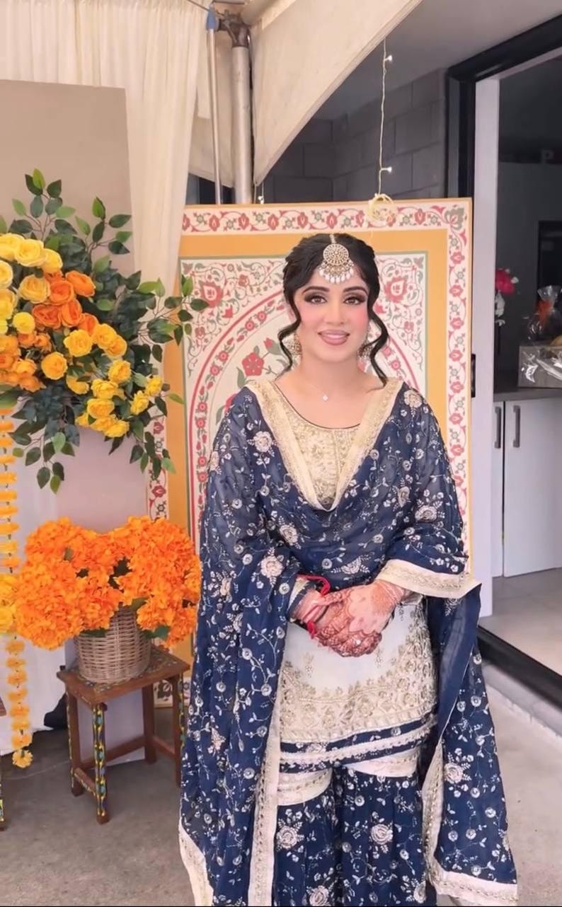 May include: A woman in a navy blue and gold embroidered outfit, including a headpiece, stands in front of a decorative backdrop. The outfit features floral patterns and intricate detailing. Floral arrangements in yellow and orange are visible.