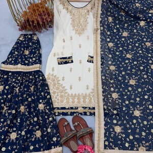 May include: An ivory-colored embroidered tunic and matching navy blue skirt and scarf set. The set features gold floral embroidery and embellishments. Brown sandals with decorative straps and pink flowers complete the ensemble.