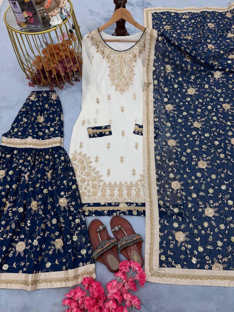 May include: An ivory-colored embroidered tunic and matching navy blue skirt and scarf set. The set features gold floral embroidery and embellishments. Brown sandals with decorative straps and pink flowers complete the ensemble.