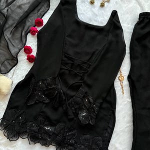 May include: Black ensemble featuring a long-sleeved top with floral embellishments on the sleeves and hem, and a lace-up back. The outfit includes matching pants and a sheer scarf. Gold earrings and a gold chain are also visible.