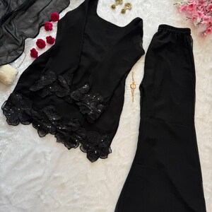 May include: Black two-piece outfit featuring a long-sleeved top and flared pants. The top has floral lace detailing along the hem and sleeves. A sheer black scarf is draped nearby. Gold earrings and a watch are also visible.