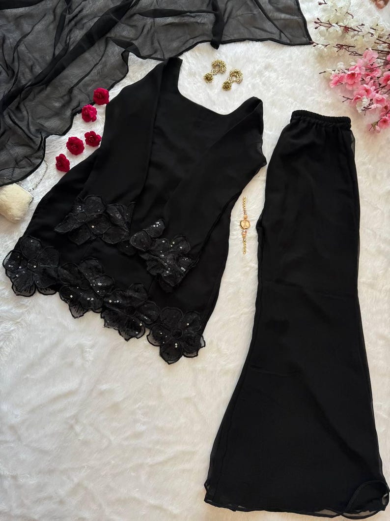 May include: Black two-piece outfit featuring a long-sleeved top and flared pants. The top has floral lace detailing along the hem and sleeves. A sheer black scarf is draped nearby. Gold earrings and a watch are also visible.