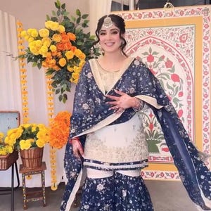 May include: A woman wearing a traditional South Asian outfit. The ensemble includes a cream-colored tunic and matching pants, overlaid with a navy blue floral patterned shawl and matching wide-leg pants. A silver headpiece completes the look.