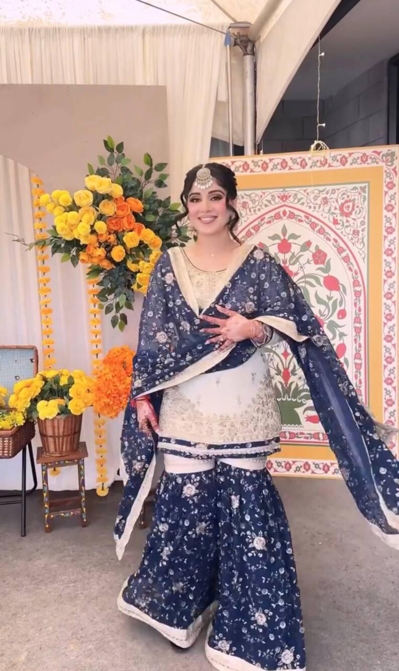 May include: A woman wearing a traditional South Asian outfit. The ensemble includes a cream-colored tunic and matching pants, overlaid with a navy blue floral patterned shawl and matching wide-leg pants. A silver headpiece completes the look.