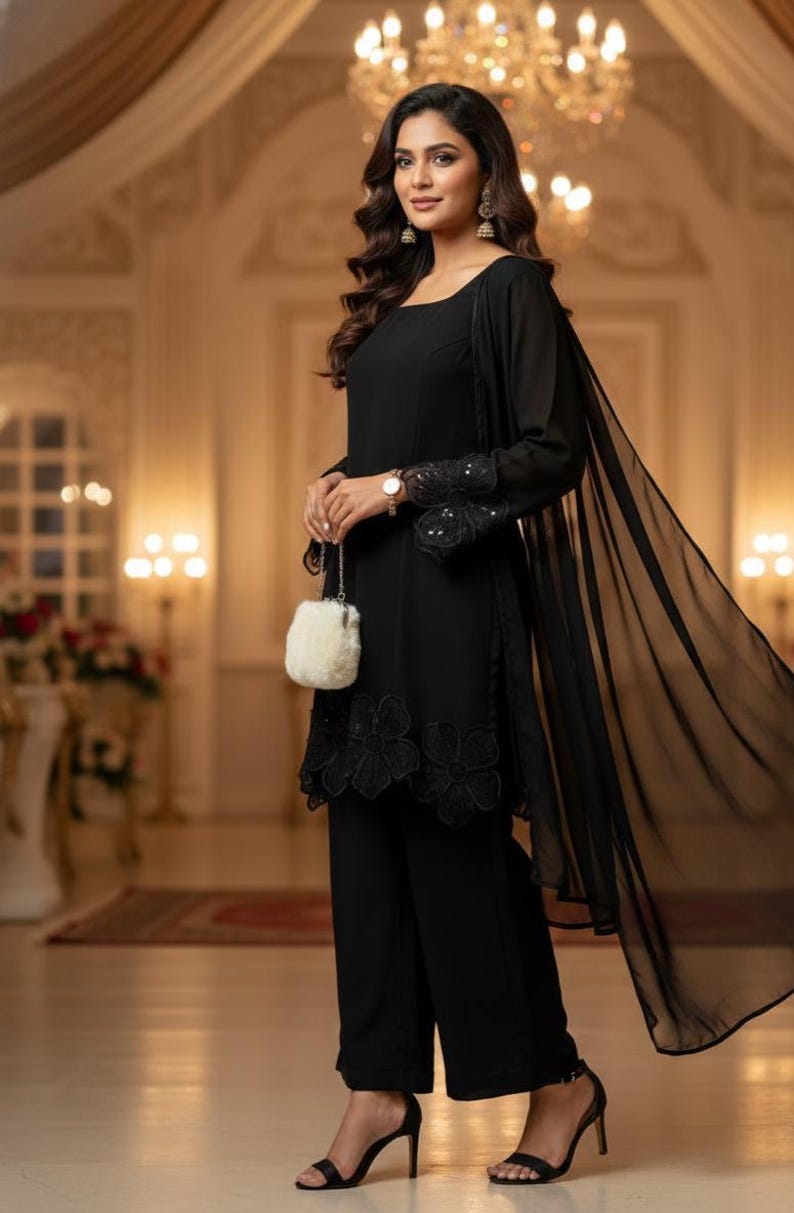 May include: A woman's black ensemble featuring a long-sleeved top with floral embroidery, matching wide-leg pants, and a sheer shawl. The outfit includes a small, fluffy white purse and black high-heeled sandals. The background is a softly lit interior.
