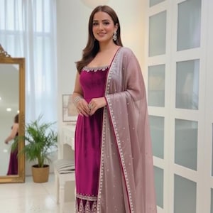 May include: A magenta velvet salwar kameez ensemble. The sleeveless top and straight-leg pants are embellished with silver accents. A sheer, light mauve dupatta with beaded trim completes the outfit. The woman is wearing silver earrings and bracelets.