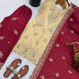 Puede incluir: Se muestra un conjunto de ropa que incluye una t&uacute;nica crema bordada, pantalones burdeos y una bufanda. La t&uacute;nica presenta intrincados bordados dorados y rojos. Tambi&eacute;n hay sandalias de cuero marr&oacute;n con detalles plateados.