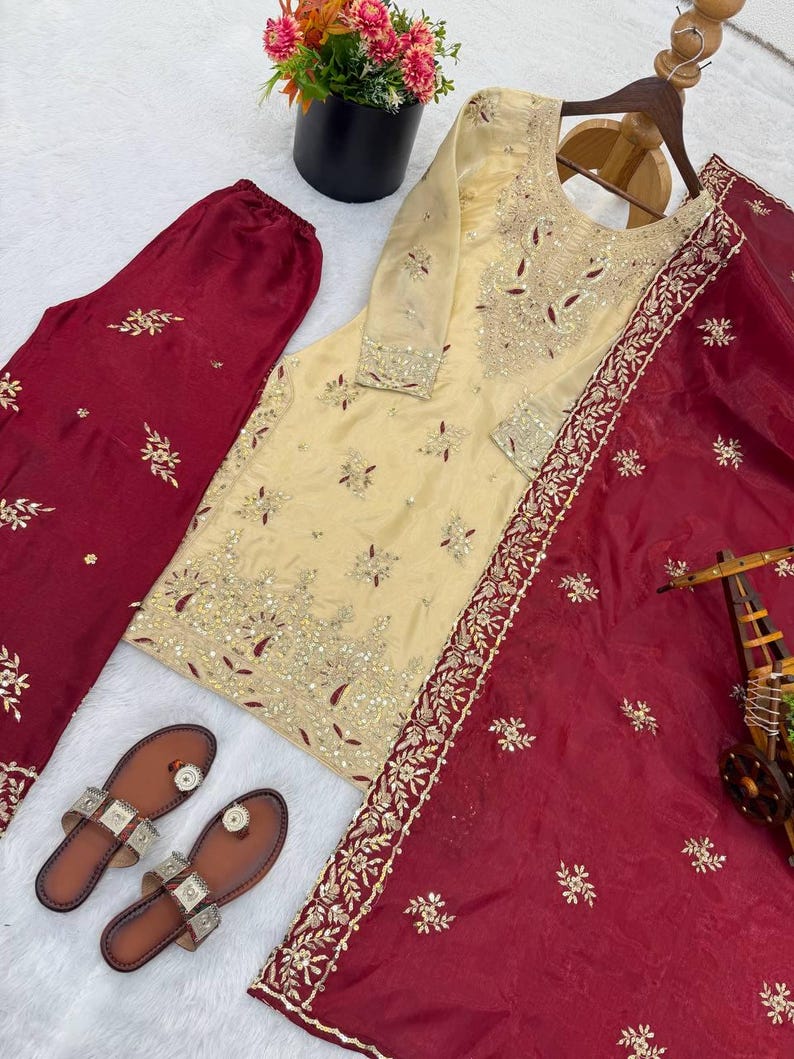 Puede incluir: Se muestra un conjunto de ropa que incluye una t&uacute;nica crema bordada, pantalones burdeos y una bufanda. La t&uacute;nica presenta intrincados bordados dorados y rojos. Tambi&eacute;n hay sandalias de cuero marr&oacute;n con detalles plateados.
