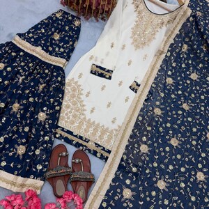 May include: An elegant ensemble featuring a white tunic with gold embroidery and navy blue bottoms with matching floral patterns. The outfit includes a matching scarf and sandals, perfect for special occasions.