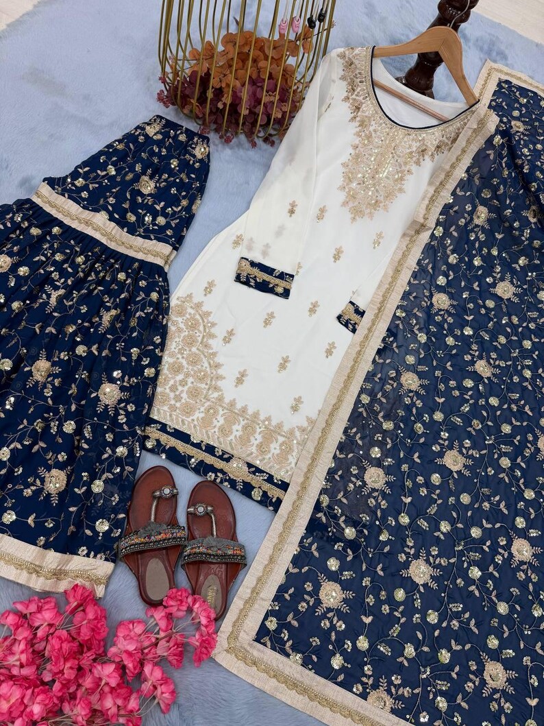 May include: An elegant ensemble featuring a white tunic with gold embroidery and navy blue bottoms with matching floral patterns. The outfit includes a matching scarf and sandals, perfect for special occasions.
