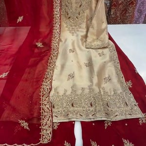 Puede incluir: Un elegante conjunto bordado de color crema con un dupatta y pantalones palazzo rojos a juego. El conjunto presenta intrincados bordados y adornos dorados, perfecto para ocasiones especiales.