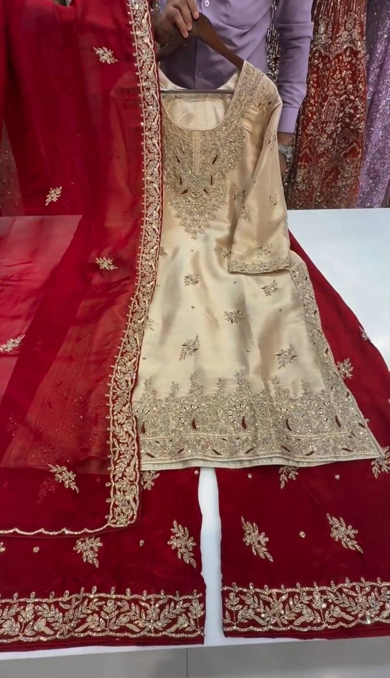 Puede incluir: Un elegante conjunto bordado de color crema con un dupatta y pantalones palazzo rojos a juego. El conjunto presenta intrincados bordados y adornos dorados, perfecto para ocasiones especiales.