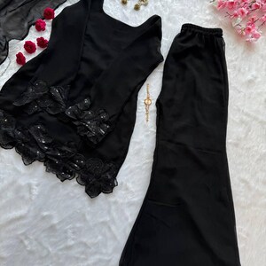 May include: Black two-piece outfit with long sleeves and flared pants. The top features floral lace detailing with sequins. A matching black scarf, gold earrings, and a gold watch are also visible.