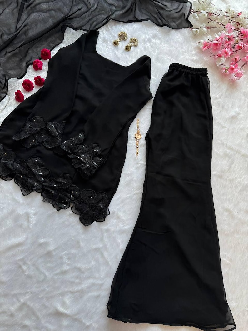 May include: Black two-piece outfit with long sleeves and flared pants. The top features floral lace detailing with sequins. A matching black scarf, gold earrings, and a gold watch are also visible.